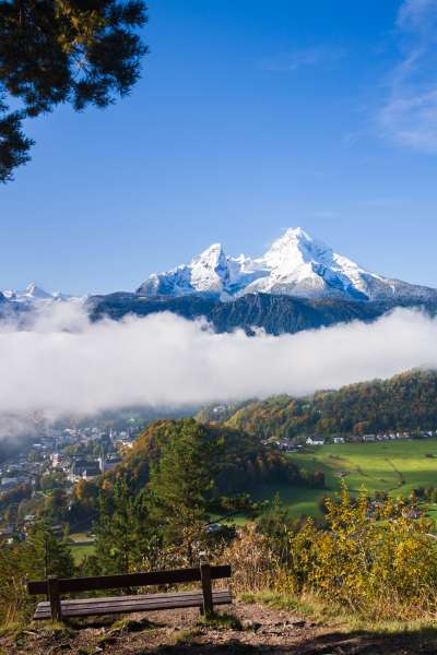 Blick von der Marxenhöhe auf den Watzmann mit schneebedeckten Gipfeln, die über einer Nebelschicht aufragen, darunter herbstliche Wälder, grüne Almwiesen und eine Holzbank im Vordergrund