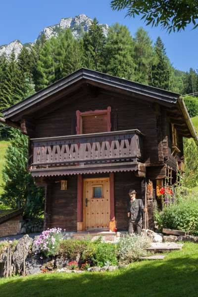 Traditionelles Almhütten-Ferienhaus am Vorderbrand mit dunklem Holz, geschnitztem Balkon und Blumengarten, vor alpiner Bergkulisse mit grünen Wäldern und Felsengipfeln