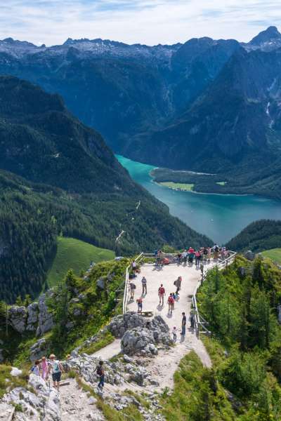 Panoramablick vom Jenner auf den türkisblauen Königssee, umgeben von bewaldeten Berghängen und schroffen Gipfeln, mit Wanderern auf dem Aussichtspunkt im Vordergrund
