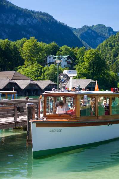 Ausflugsboot am Anleger in Schönau am Königssee mit Passagieren, im Hintergrund traditionelle Gebäude und bewaldete Berghänge