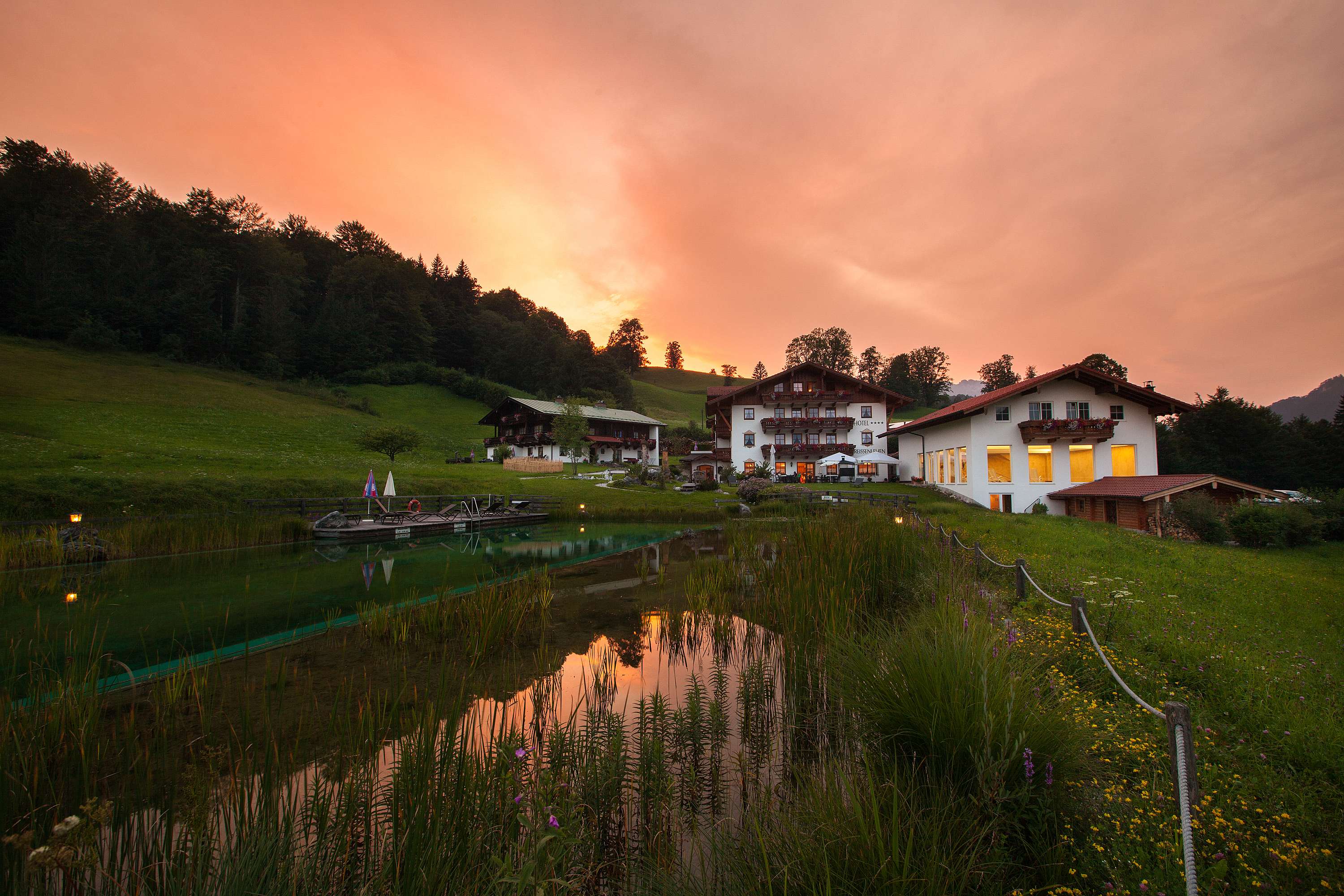 Hotel Reissenlehen OHG in Bischofswiesen bei Abenddämmerung mit dramatischem orangefarbenem Himmel, Hotelgebäuden am Hang und Naturteich im Vordergrund