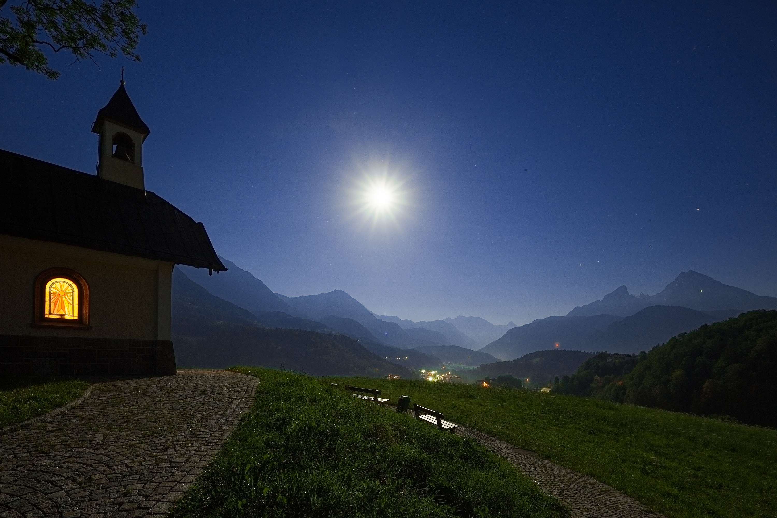 Bergkapelle Lockstein bei Nacht mit beleuchtetem Fenster, Vollmond am Sternenhimmel und Blick ins beleuchtete Tal mit Bergsilhouetten